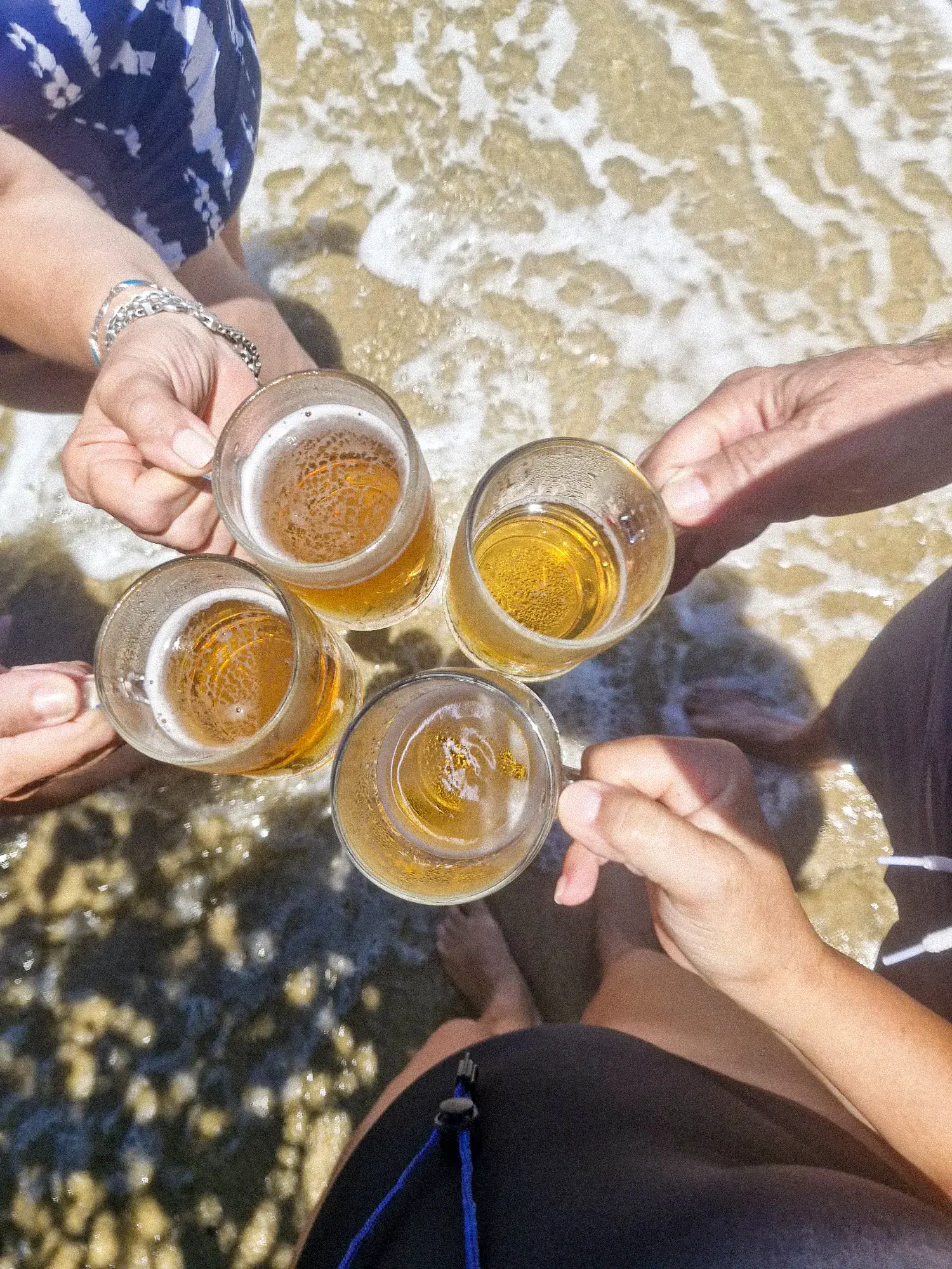 Cuatro manos brindando con tragos en la orilla de una playa, al atardecer