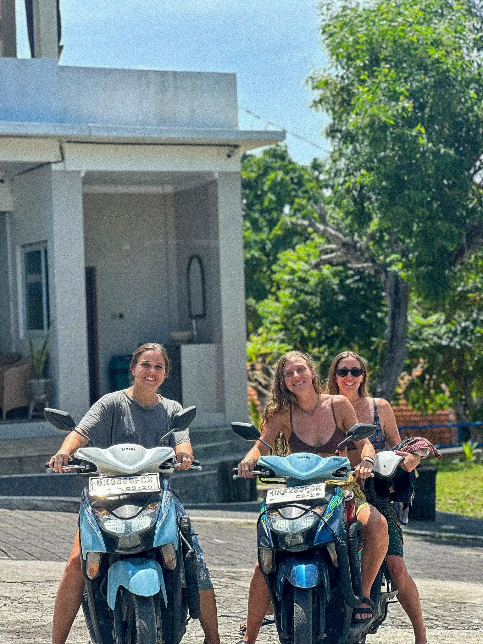 Tres viajeras en dos motos, sonriendo, en una calle de Bali