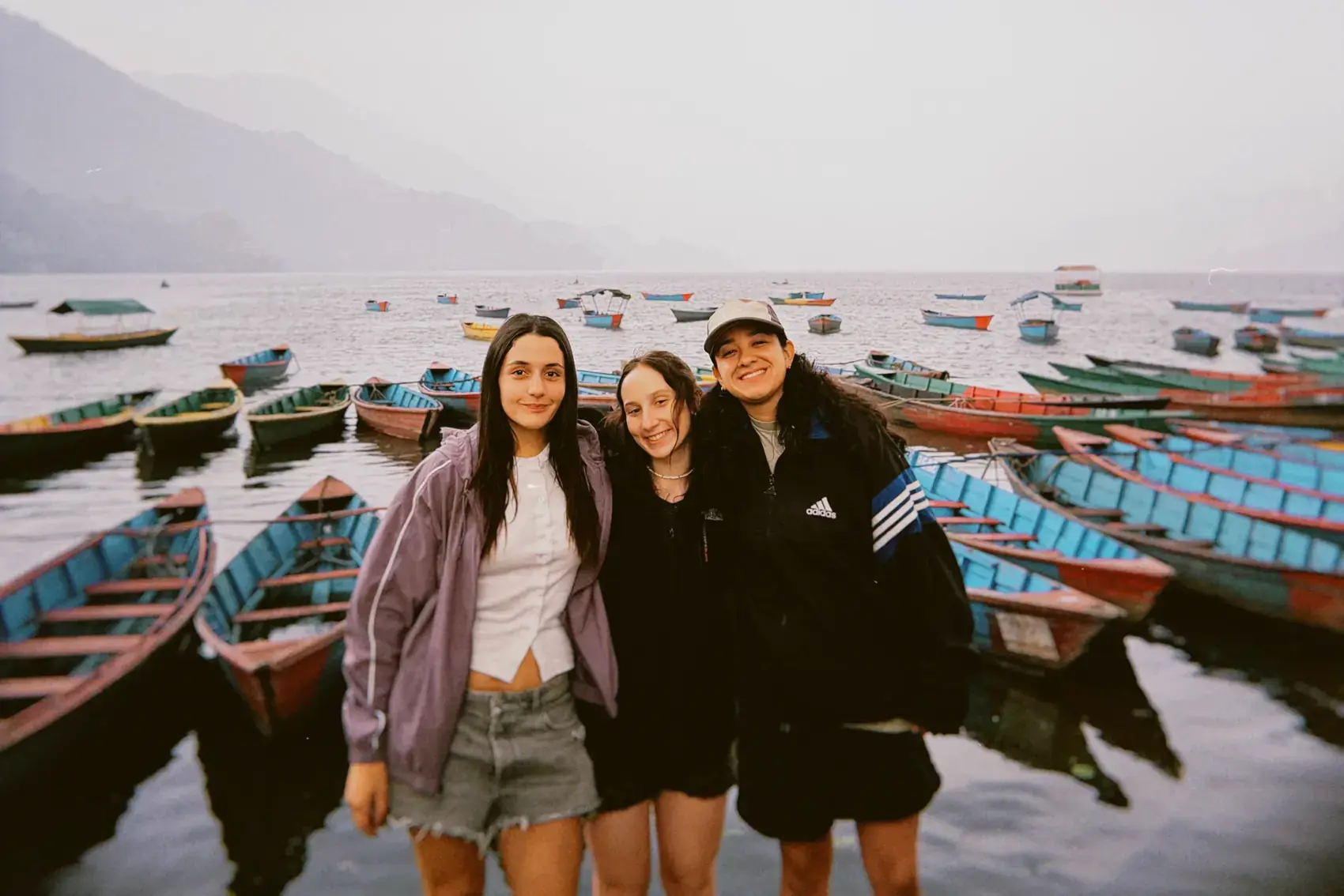 Tres viajeras abrazadas en el muelle del lago Phewa, con los botes de colores atrás