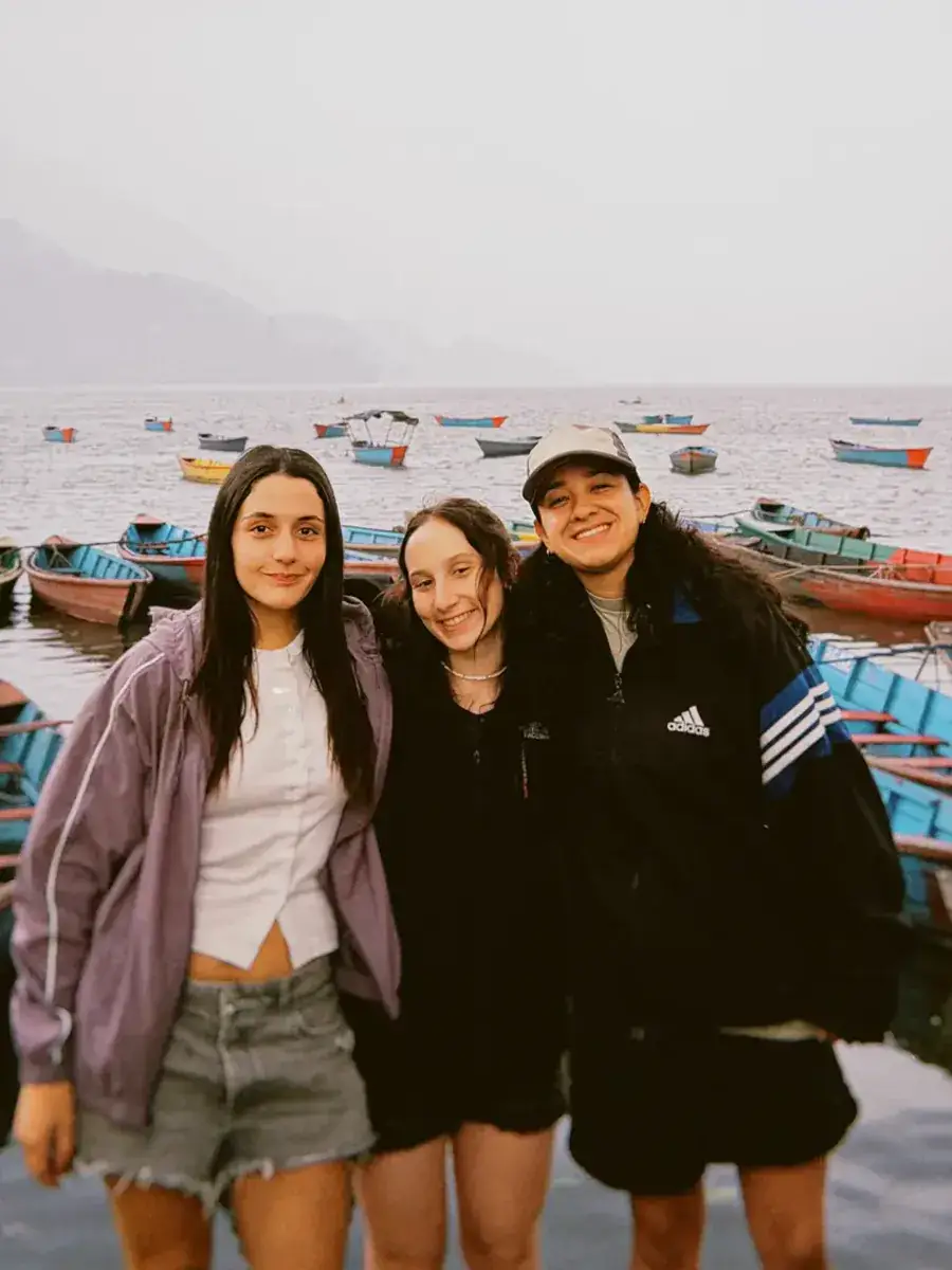 Tres viajeras abrazadas en el muelle del lago Phewa, con los botes de colores atrás