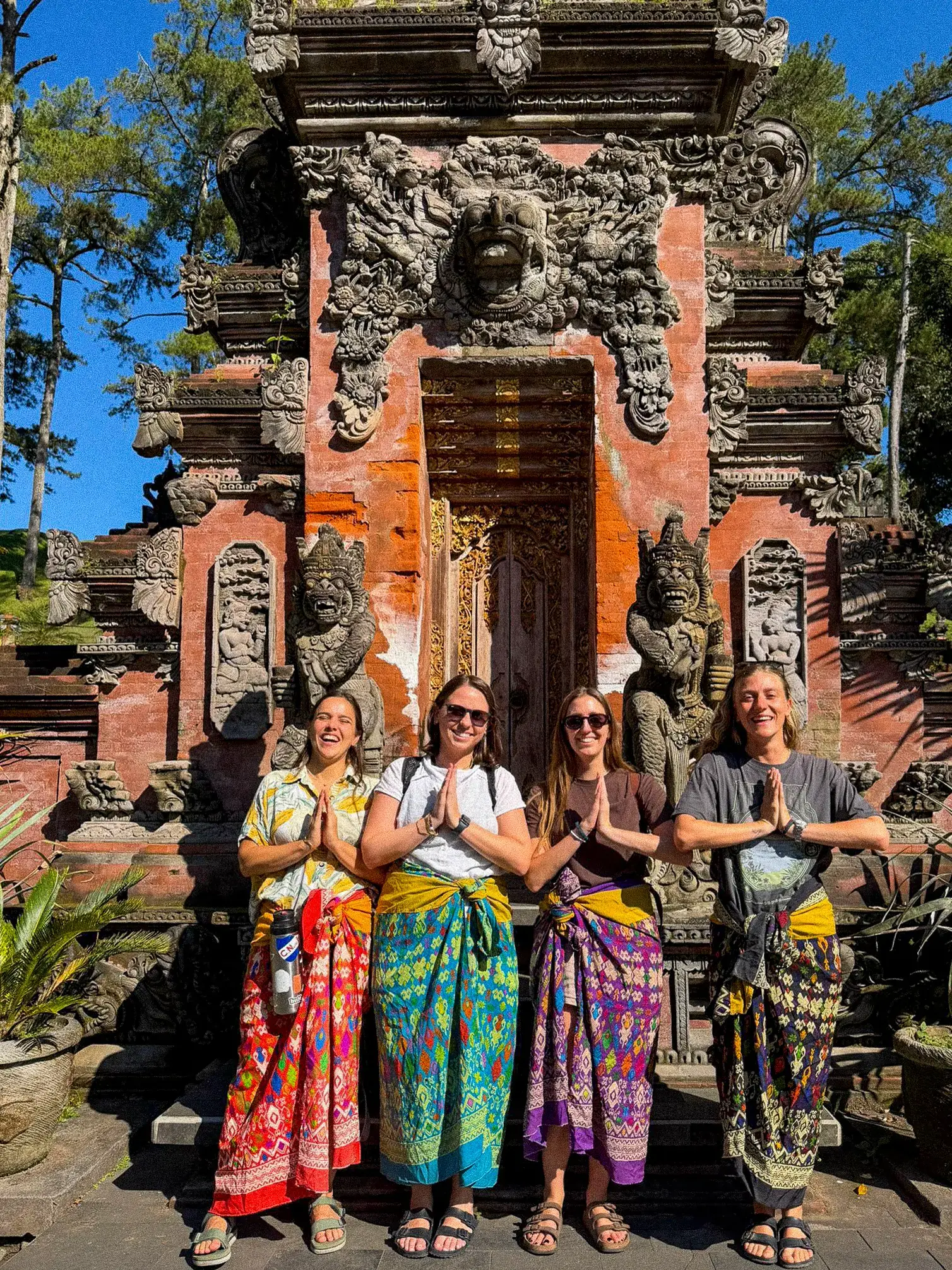 Cuatro viajeras con sarong tradicional frente a un templo balinés, con las manos juntas en gesto de oración