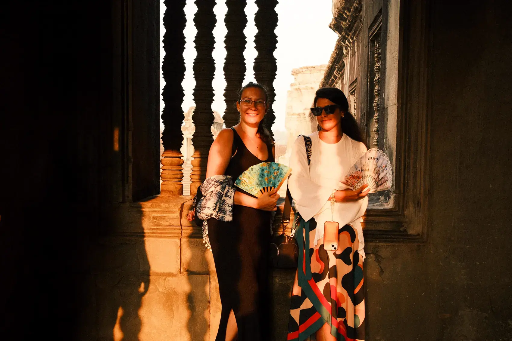 Dos viajeras con abanicos, paradas junto a una ventana tallada de Angkor Wat, con luz dorada
